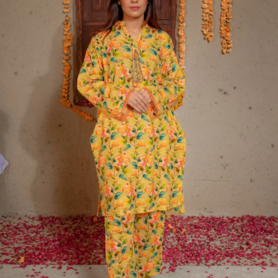 A girl standing indoors wearing a yellow floral lace outfit and tan sandals. Flower petals are scattered on the floor, and marigold garlands hang on the wall.